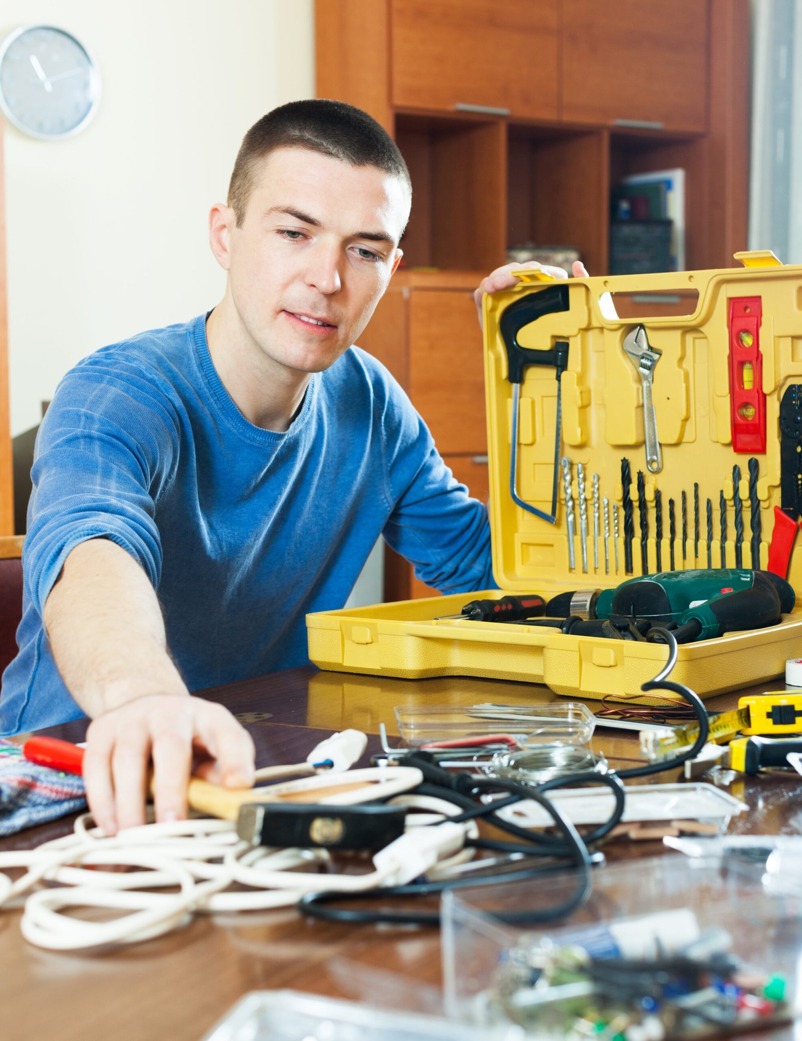 Young handsome man sitting by table with toolbox and reaching to get hammer